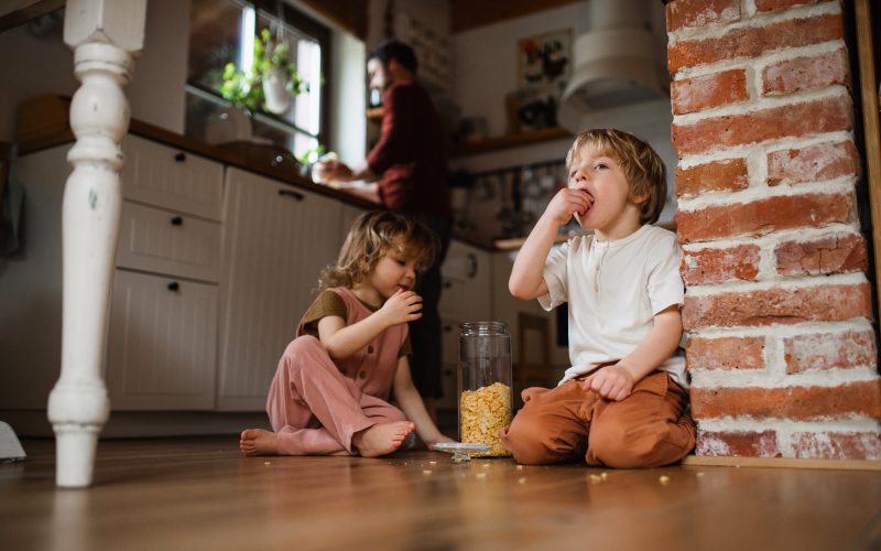 Two small children with father indoors at home, eating cornflakes on floor